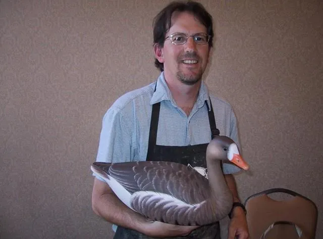 A man in a blue shirt and apron proudly holds a realistic decoy goose, showcasing craftsmanship in wildlife art.