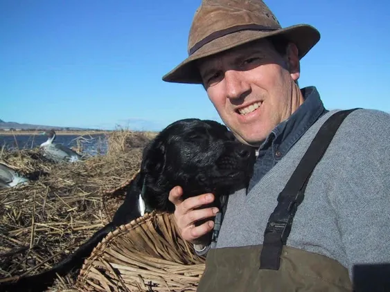 A man in a hat poses for a selfie with a black dog by a wetlands area, showcasing a bond between them amidst a natural landscape.