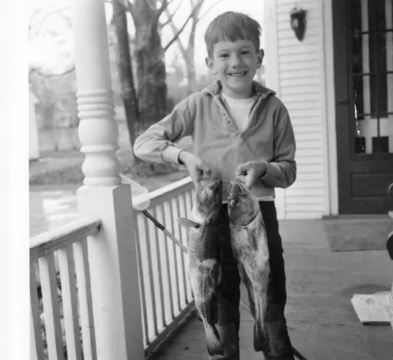 Young boy proudly holding two large fish on a porch, smiling for the camera. The scene captures a moment of childhood joy and fishing achievement.