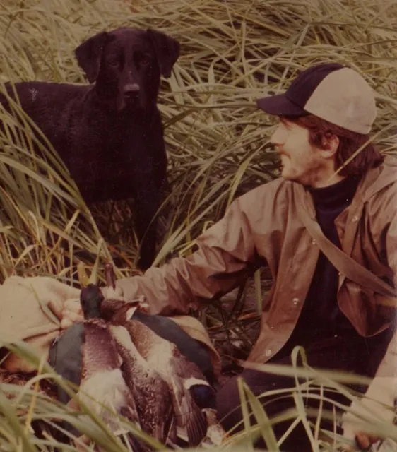 A man sits in tall grass next to a pile of ducks, with a black Labrador Retriever nearby, capturing a moment of a successful hunting trip.