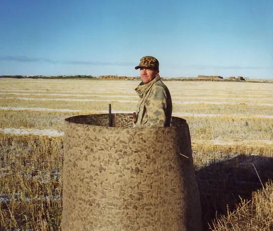 A hunter stands inside a camouflage blind in an open field, preparing for the hunt in a tranquil, expansive landscape.
