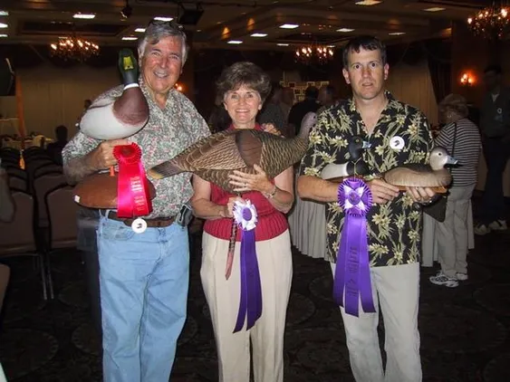 Three individuals proudly display their award-winning duck decoys at a competition, showcasing craftsmanship and competition spirit in a decorated venue.